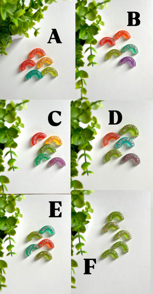 Groups of five or six translucent glittery rainbow-shaped resin pieces in various colors on a white surface near green plant leaves