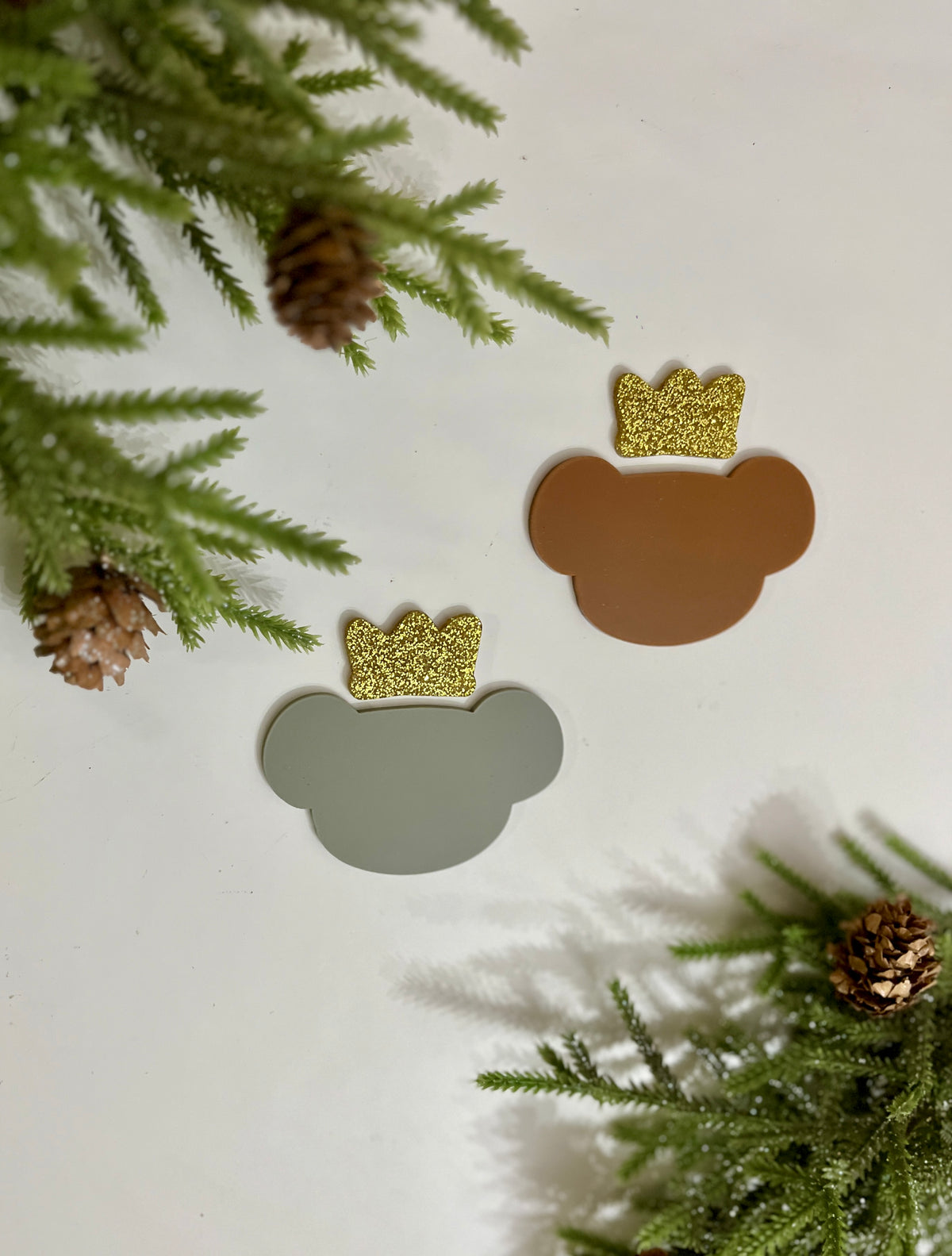 Two bear-shaped cutouts with glittery gold crowns surrounded by pine branches and cones on a white surface