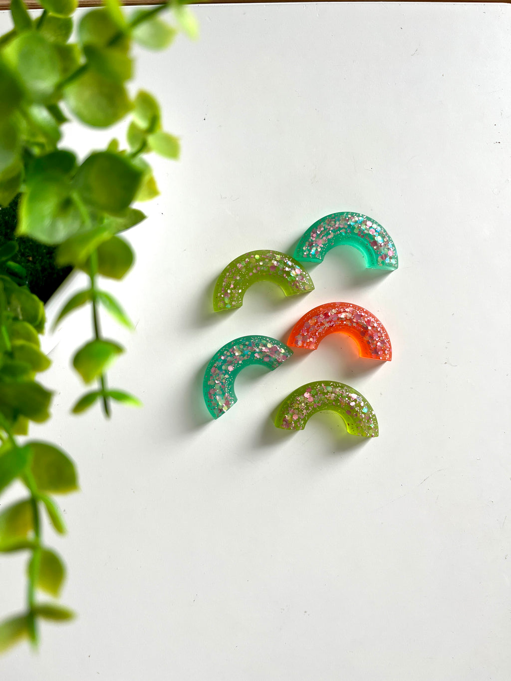 Five glitter-filled translucent resin half-circle pieces in green, teal, and orange on a white surface near green leaves
