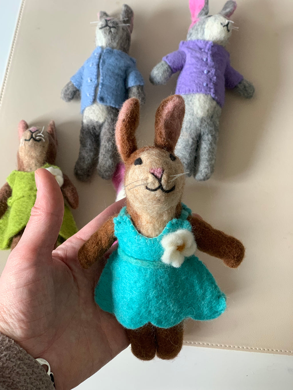 Handfelt brown bunny doll wearing a turquoise dress with a white flower held in hand with three other dolls on a beige surface