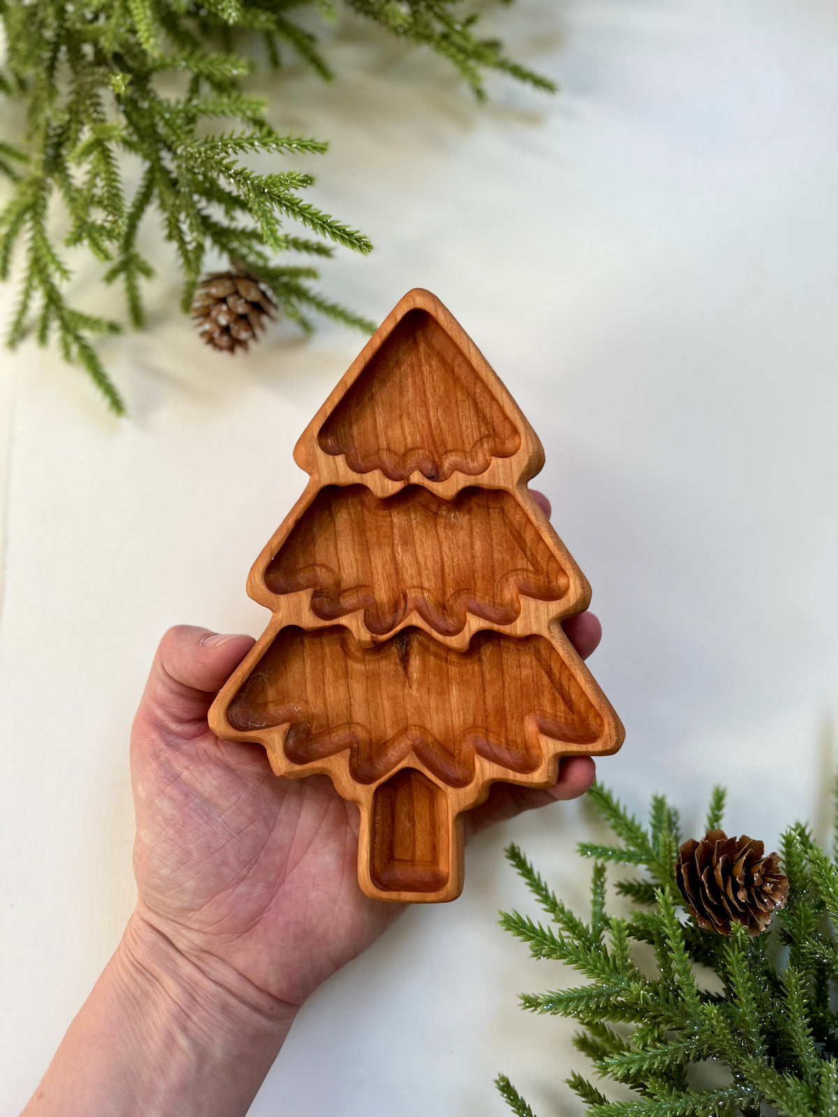 Hand holding a carved wooden tray shaped like a tiered Christmas tree with pine cones and branches nearby