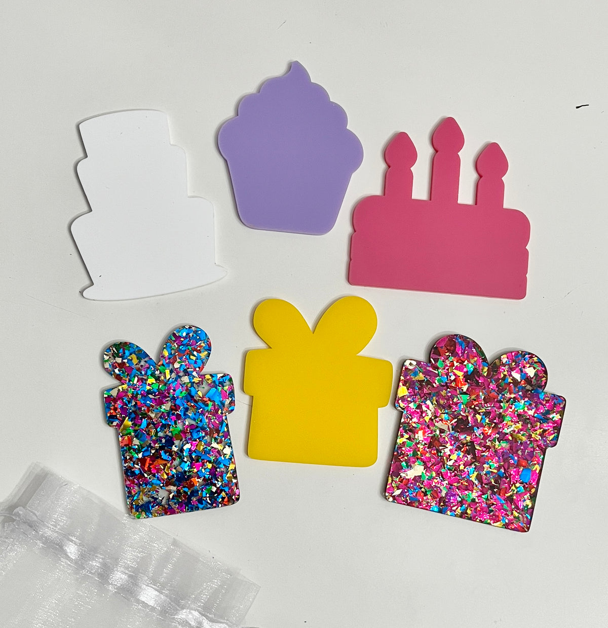 Five colorful acrylic shapes including white cake, purple cupcake, pink birthday cake with candles, and glittery blue and pink gift boxes on white background