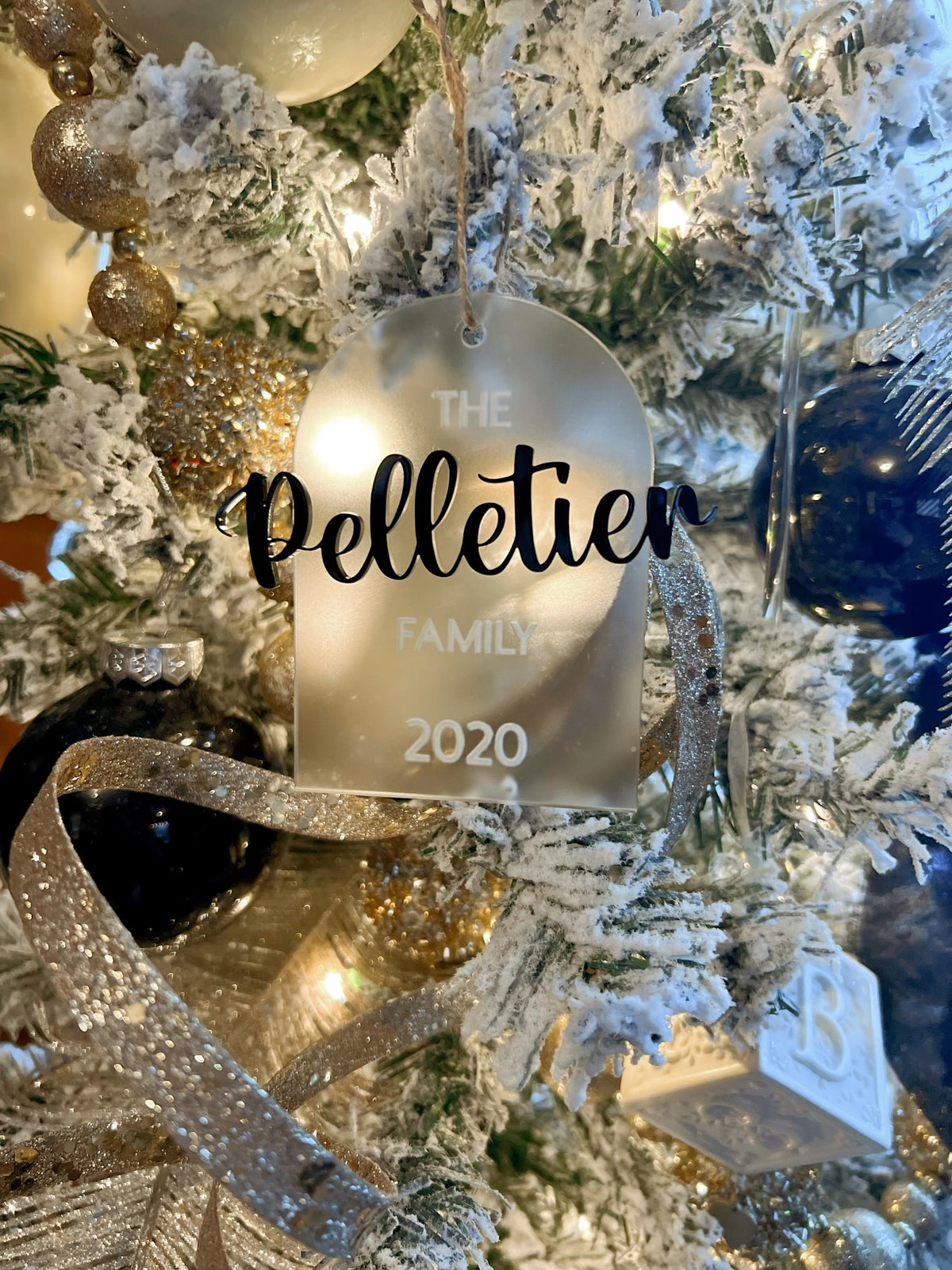 Frosted acrylic arch ornament with black and white text displaying "The Pelletier Family 2020" hanging on snowy branches