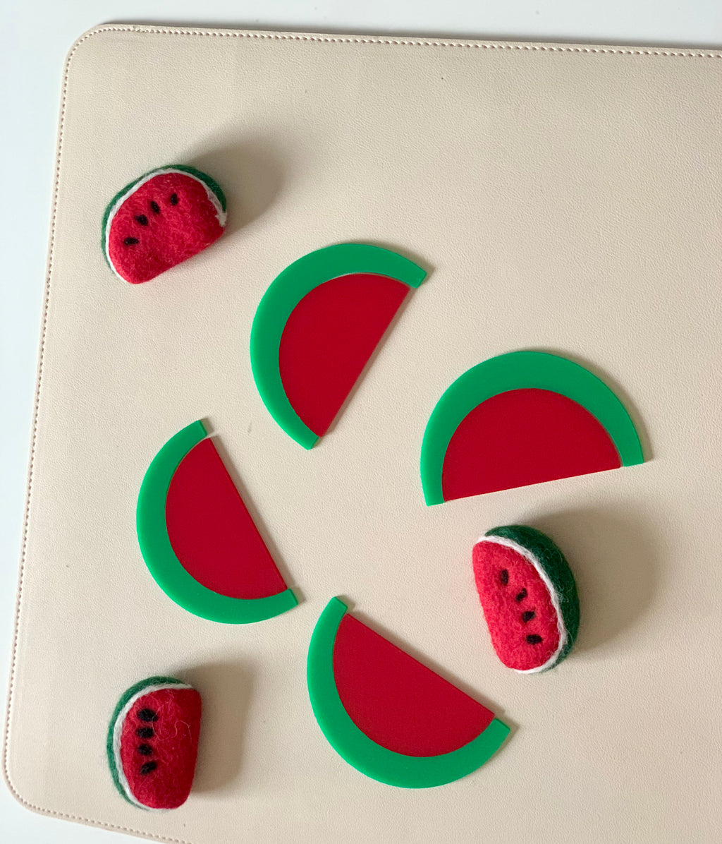 Red and green watermelon-shaped felt toys and plastic watermelon slice pieces arranged on a beige surface