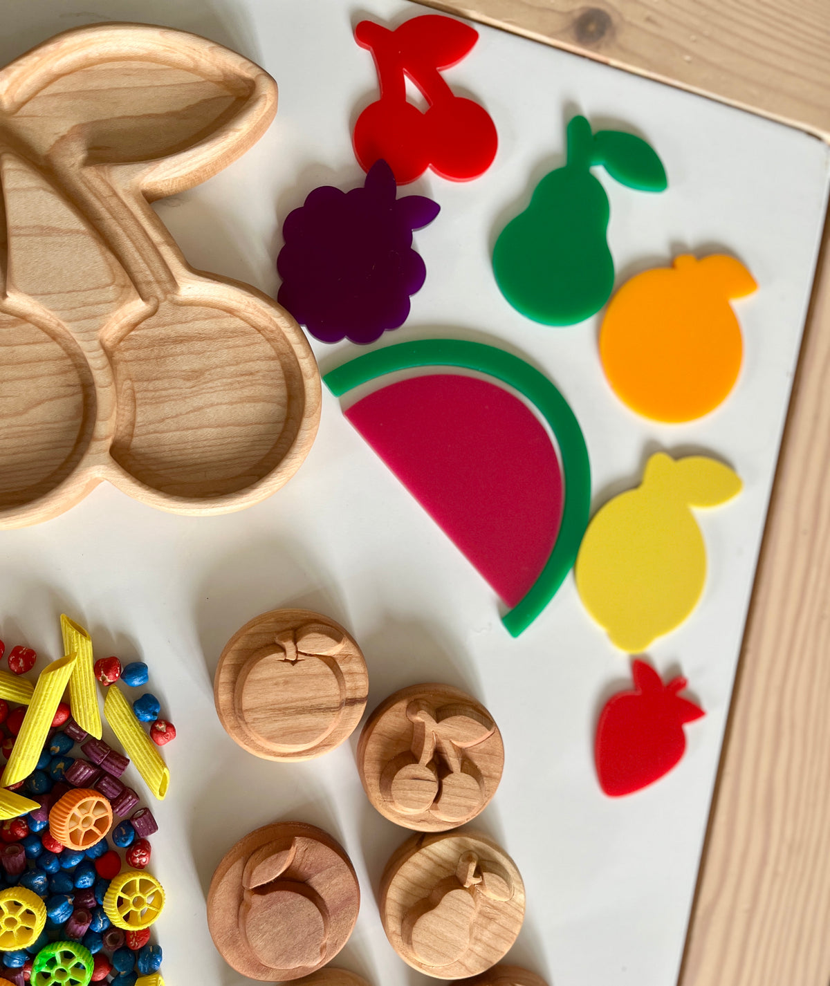 Wooden cherry-shaped tray with colorful fruit-shaped cutouts, wooden fruit stamps, and multicolored pasta pieces on white surface