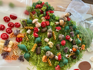 Variety of colorful dried pasta shapes, small red mushroom decorations, and acorns scattered on green textured surface
