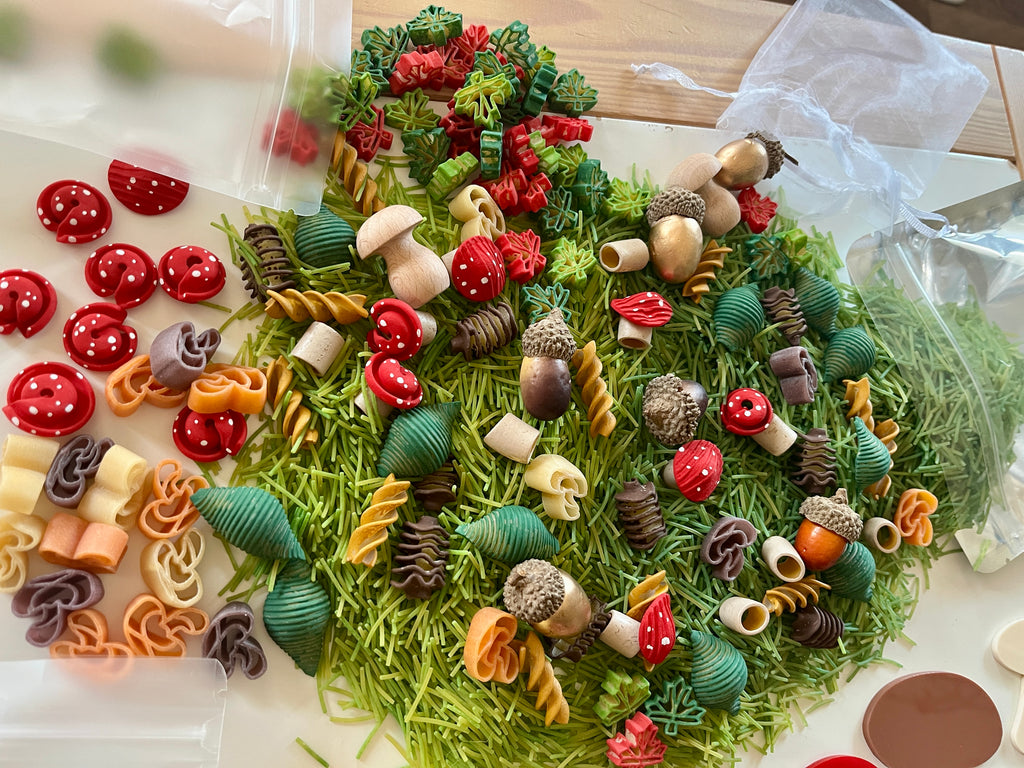 Variety of colorful dried pasta shapes, small red mushroom decorations, and acorns scattered on green textured surface