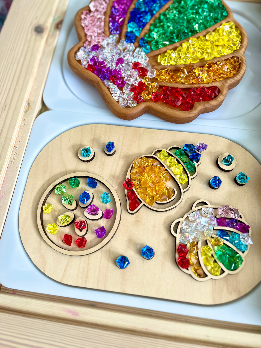 Wooden tray with colorful translucent gem beads sorted into shapes including a shell, acorn, and ladybug on white surface