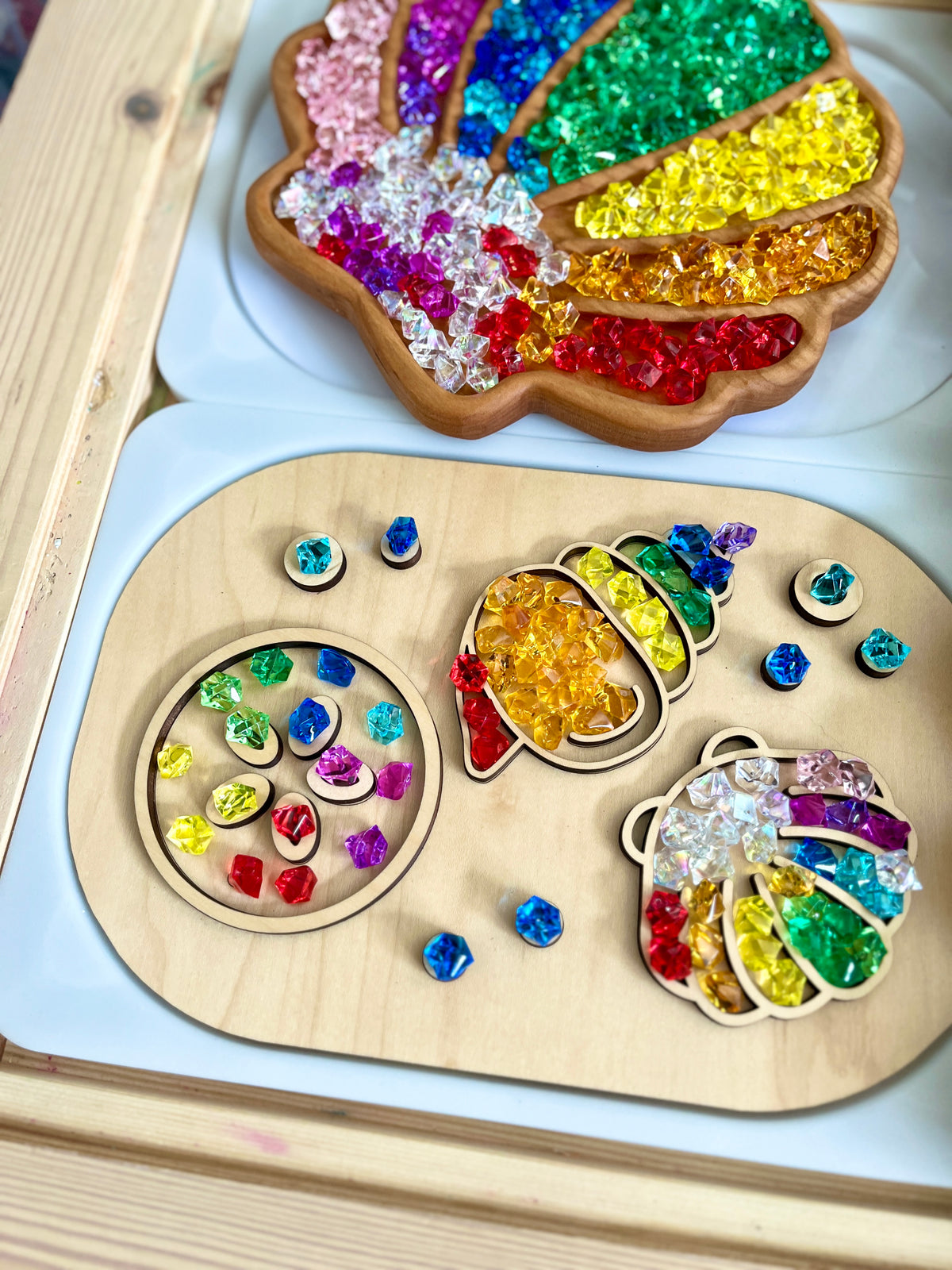 Wooden tray with colorful translucent gem beads sorted into shapes including a shell, acorn, and ladybug on white surface