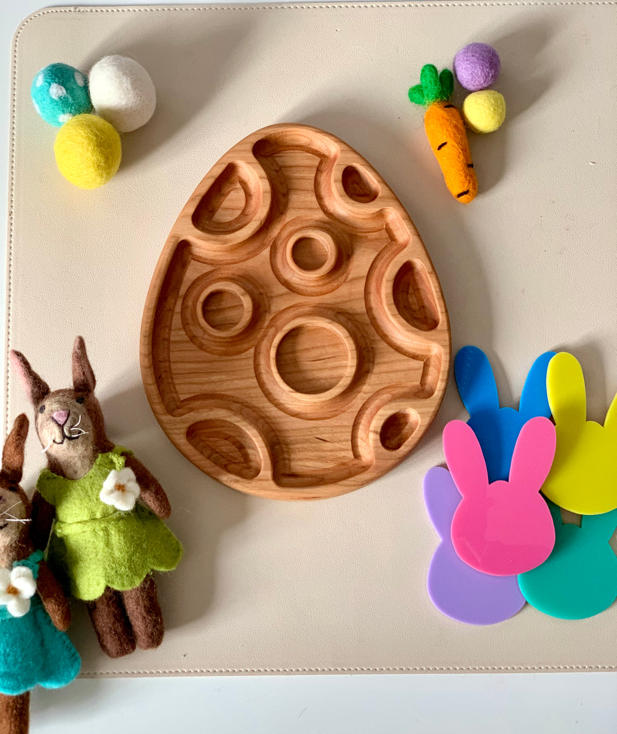 Wooden egg-shaped tray with carved circular designs surrounded by felt balls, toy carrots, bunny-shaped cutouts, and plush rabbits