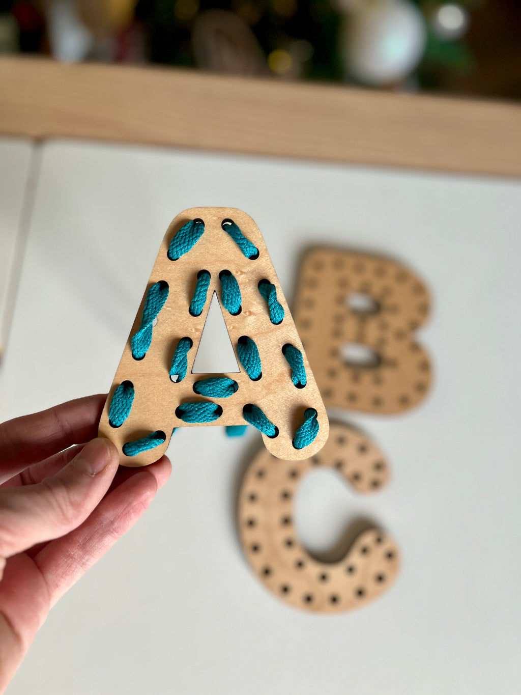 Hand holding a wooden letter A threaded with bright blue lace, with wooden letters B and C blurred in the background, Alphabet Lacing Letters (A, B, C shown — full alphabet optional), handcrafted homeschool education resources.