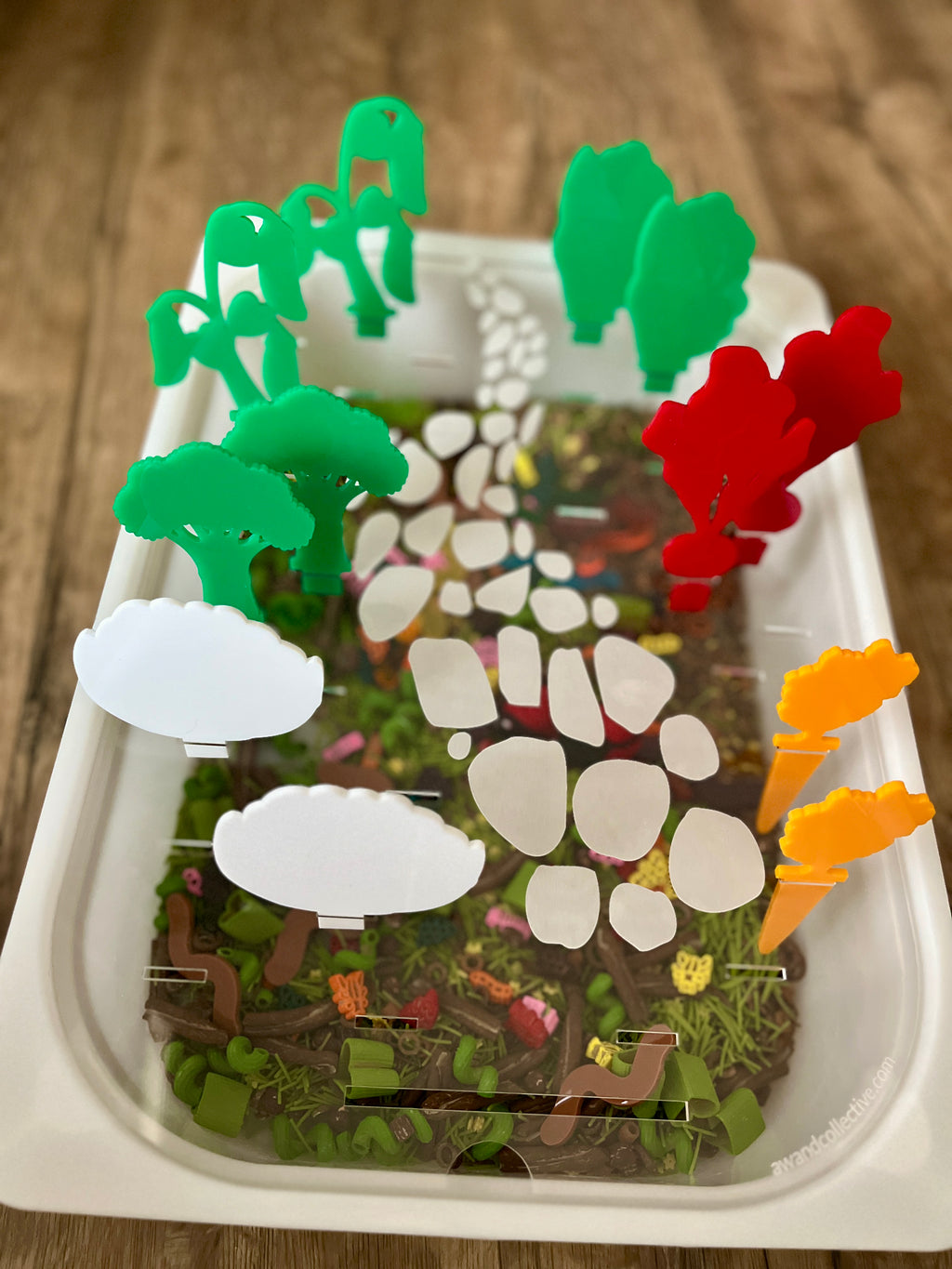 White tray with a nature-themed base design holding colorful translucent acrylic pegs in green, red, yellow, and white shapes resembling trees and bushes, featuring STEM Insert – Acrylic Insert with Different Peg Options - Neighborhood, Veggie Garden, Flower Garden, Under the Sea + More, handcrafted, homeschool, education resources.
