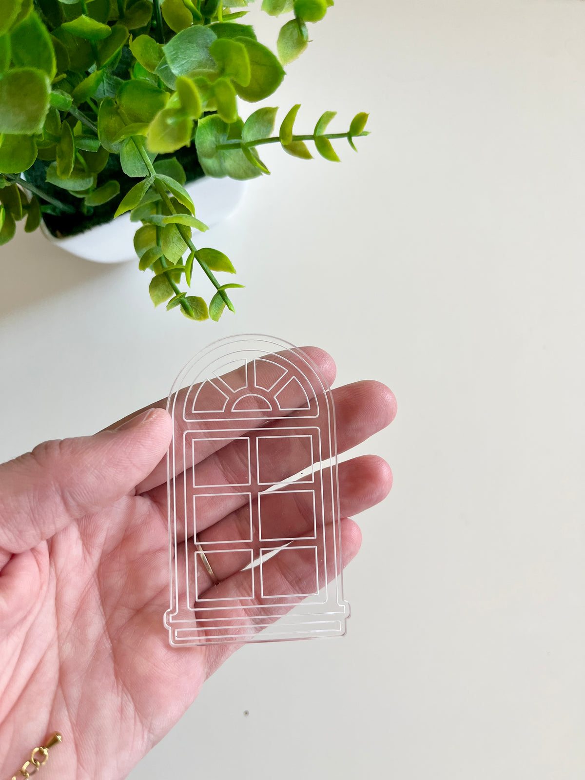 Hand holding a transparent acrylic window-shaped cutout with a small green potted plant in the background