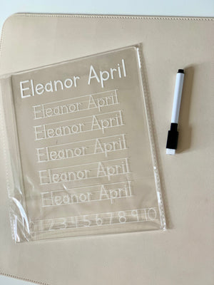 Transparent handwriting practice board with traced letters and numbers beside a black and white marker on beige surface