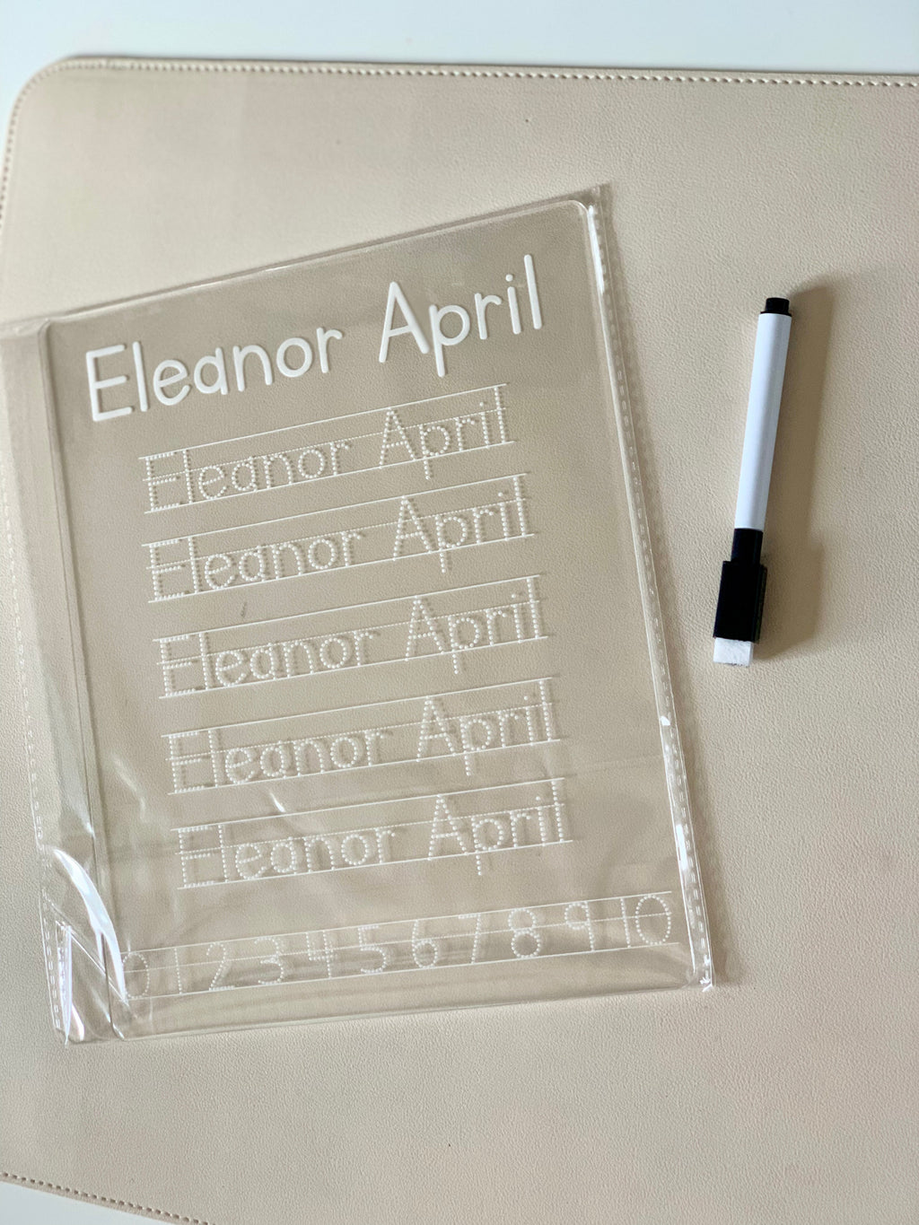 Transparent handwriting practice board with traced letters and numbers beside a black and white marker on beige surface