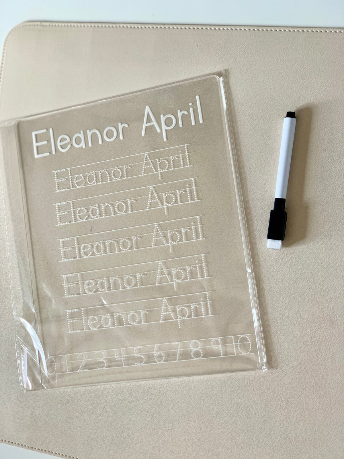 Transparent handwriting practice board with traced letters and numbers beside a black and white marker on beige surface