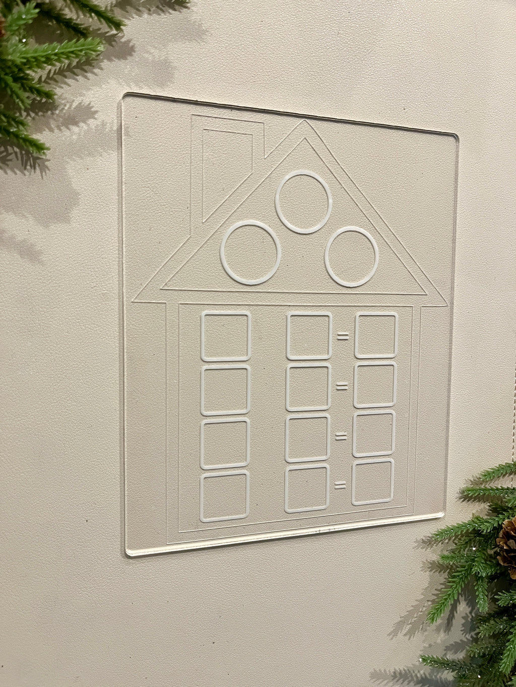 Clear acrylic board with etched house shape and geometric patterns displayed on beige surface with greenery edges
