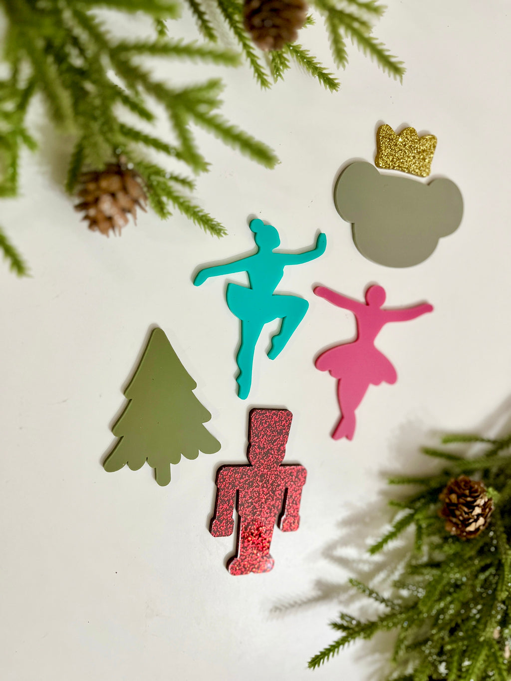 Five colorful holiday-themed cutouts including a tree, ballerinas, a nutcracker, and a bear with a gold crown near pine branches and cones