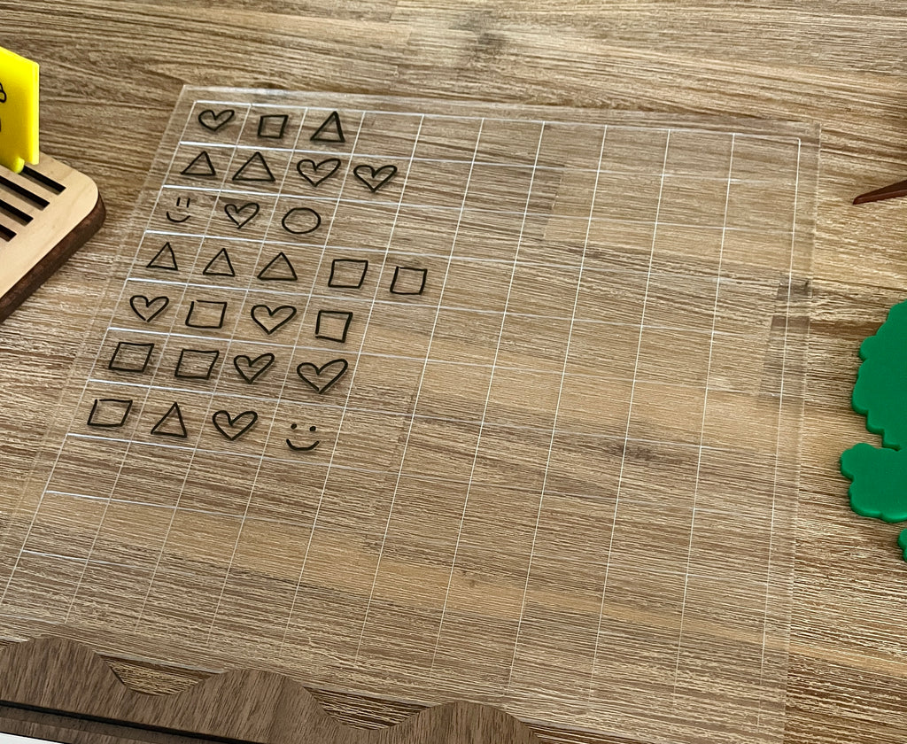 Dry Erasable Blank Open Ended 100 Frame Hundred Board on wood surface featuring transparent grid with black marker drawings of hearts, squares, triangles, and smiley face, handcrafted homeschool education resources.