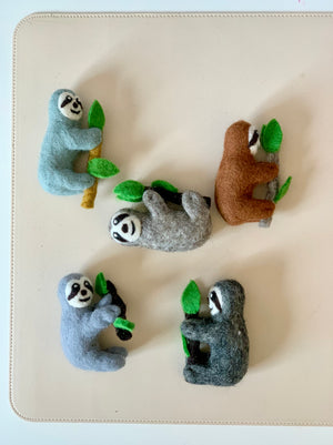 Five felt sloth figurines in various colors clinging to felt branches with green leaves on a cream background