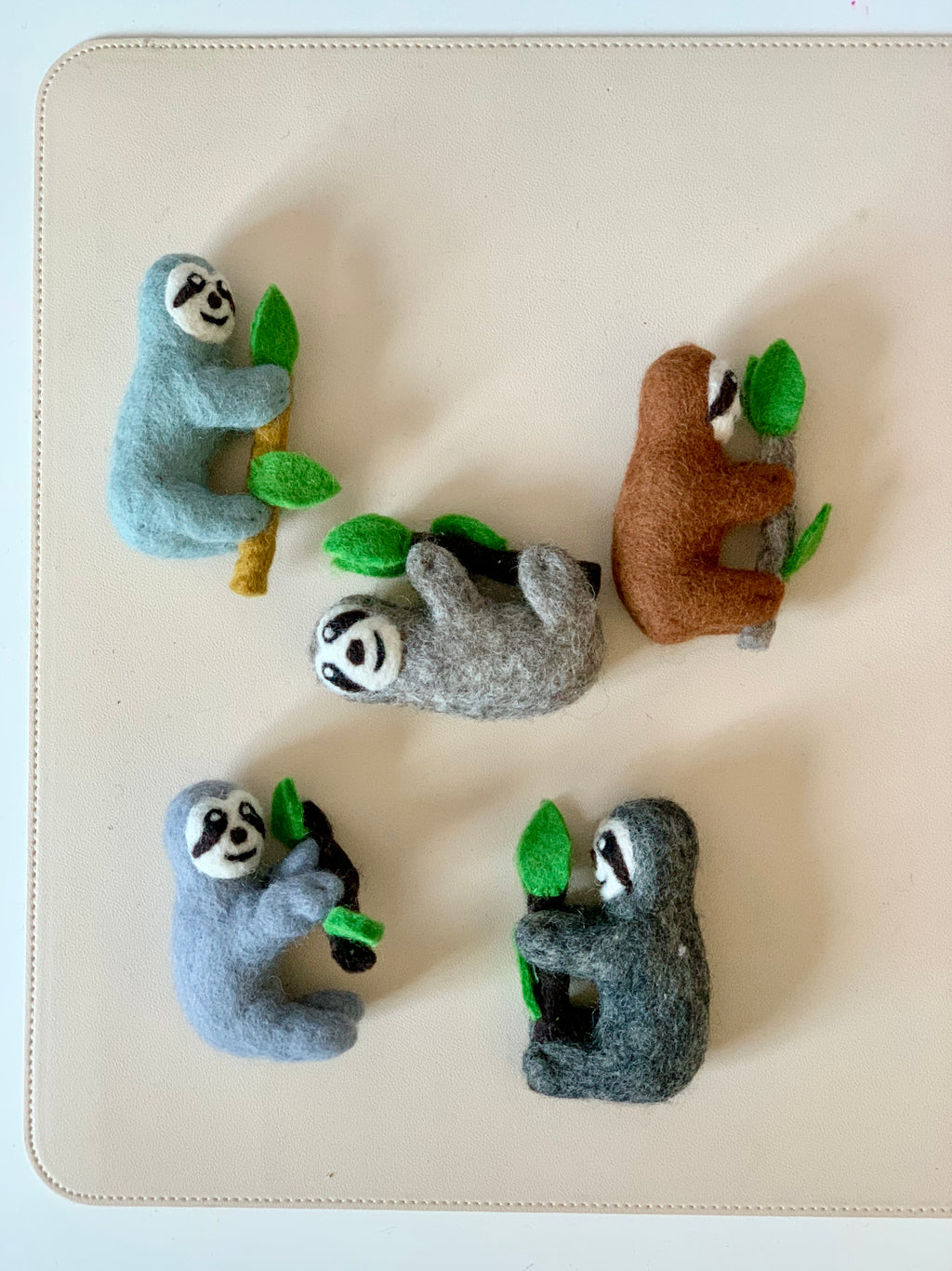 Five felt sloth figurines in various colors clinging to felt branches with green leaves on a cream background