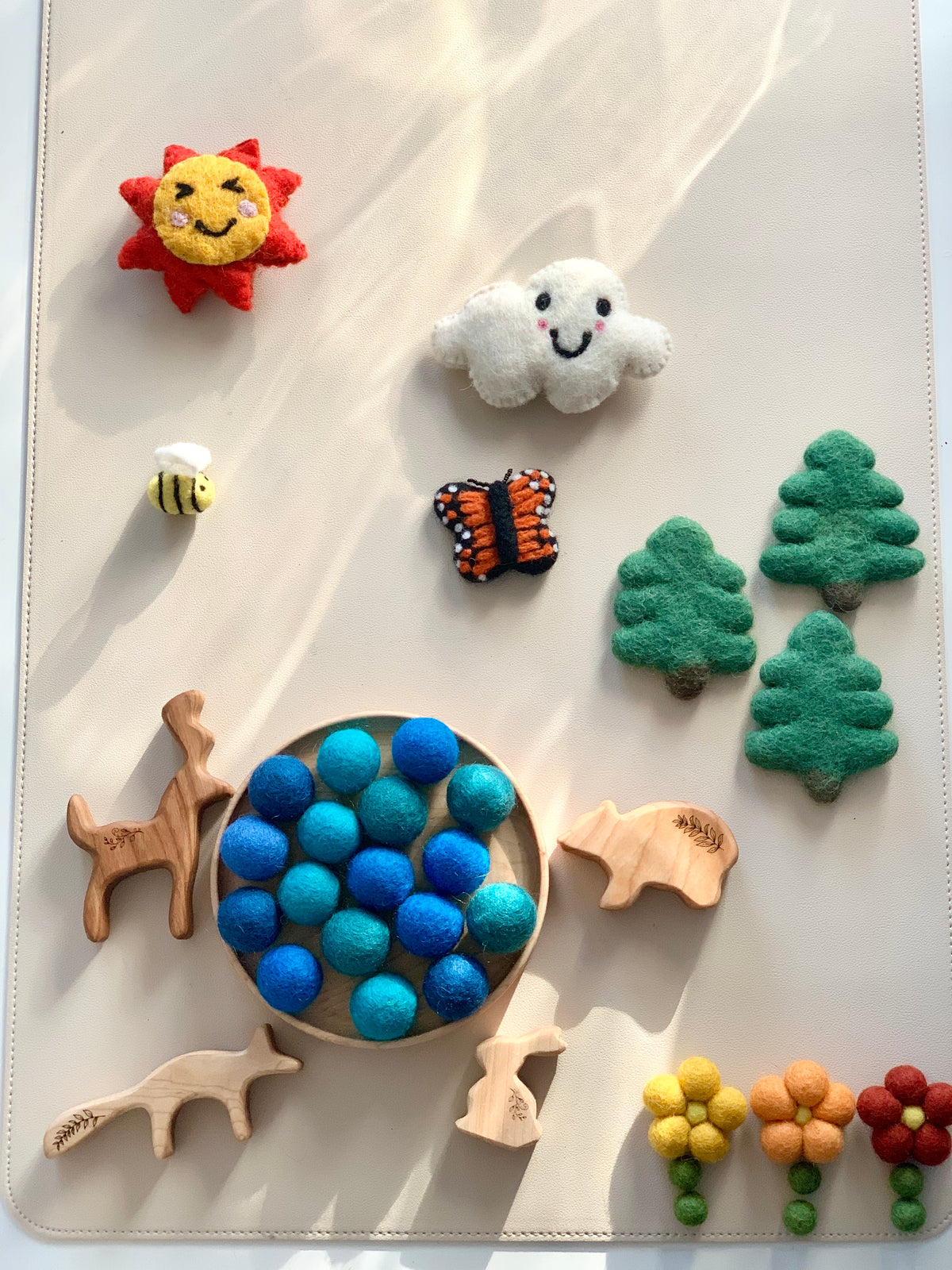 Assorted felt and wooden nature-themed pieces including sun, cloud, bee, butterfly, trees, animals, flowers, and blue felt balls in a tray
