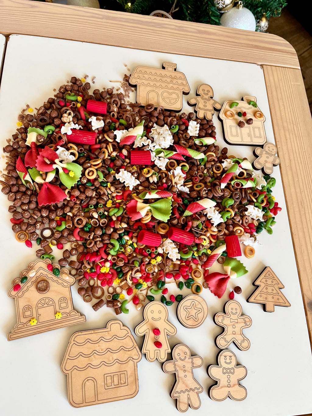 Wooden holiday-themed cutouts including gingerbread people and houses with colorful pasta and small brown cereal pieces scattered on white surface