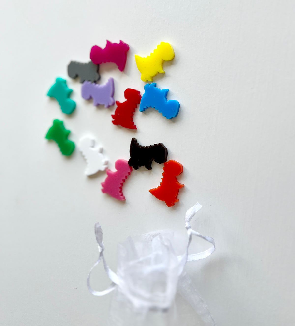Assorted colorful plastic dinosaur-shaped game pieces scattered above a small sheer white drawstring pouch on a white background