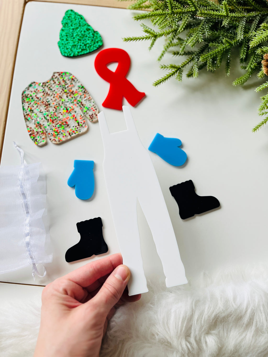 Hand holding white acrylic cutout of overalls with separate red scarf, blue mittens, black boots, green hat, and festive sweater pieces nearby