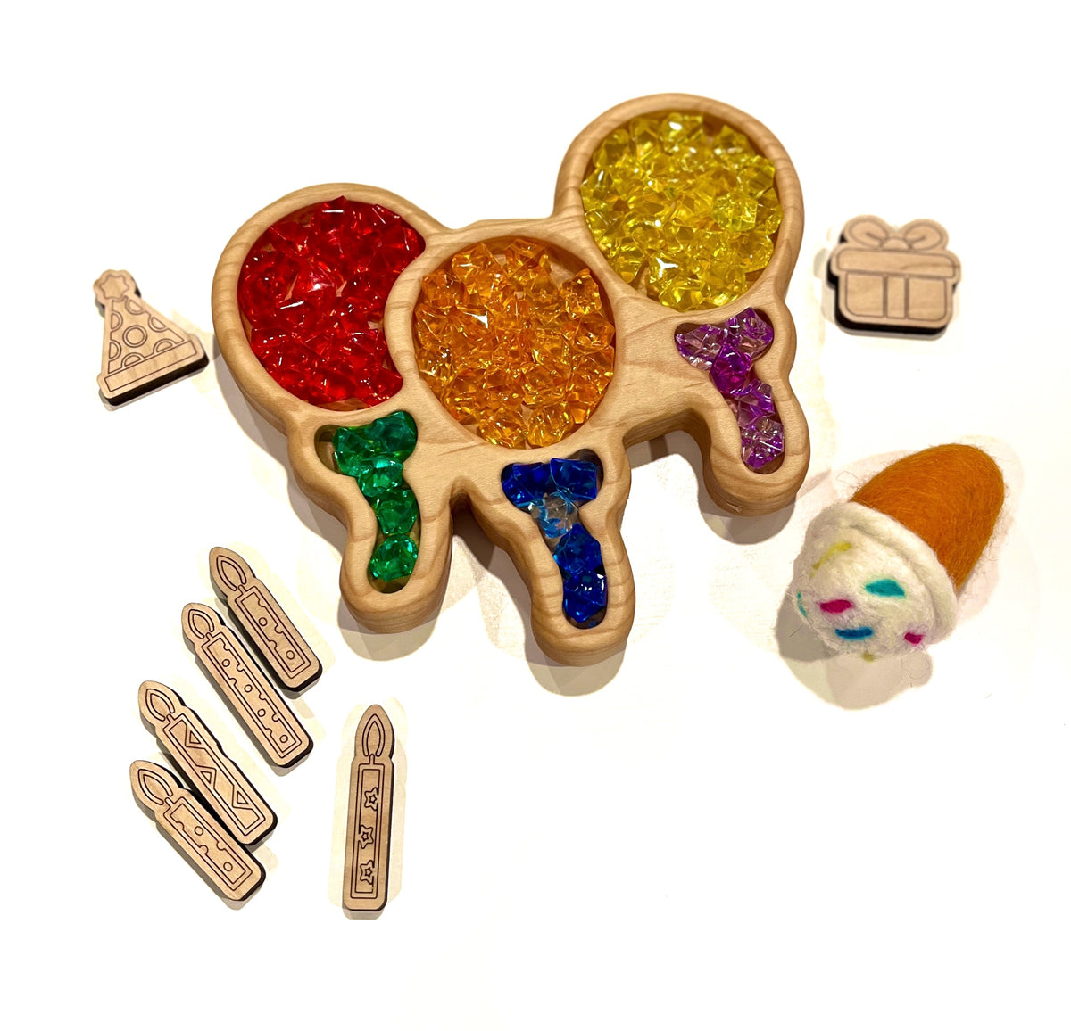 Wooden tray with colorful translucent beads shaped like balloons, surrounded by felt toy and wooden party-themed tokens on white background