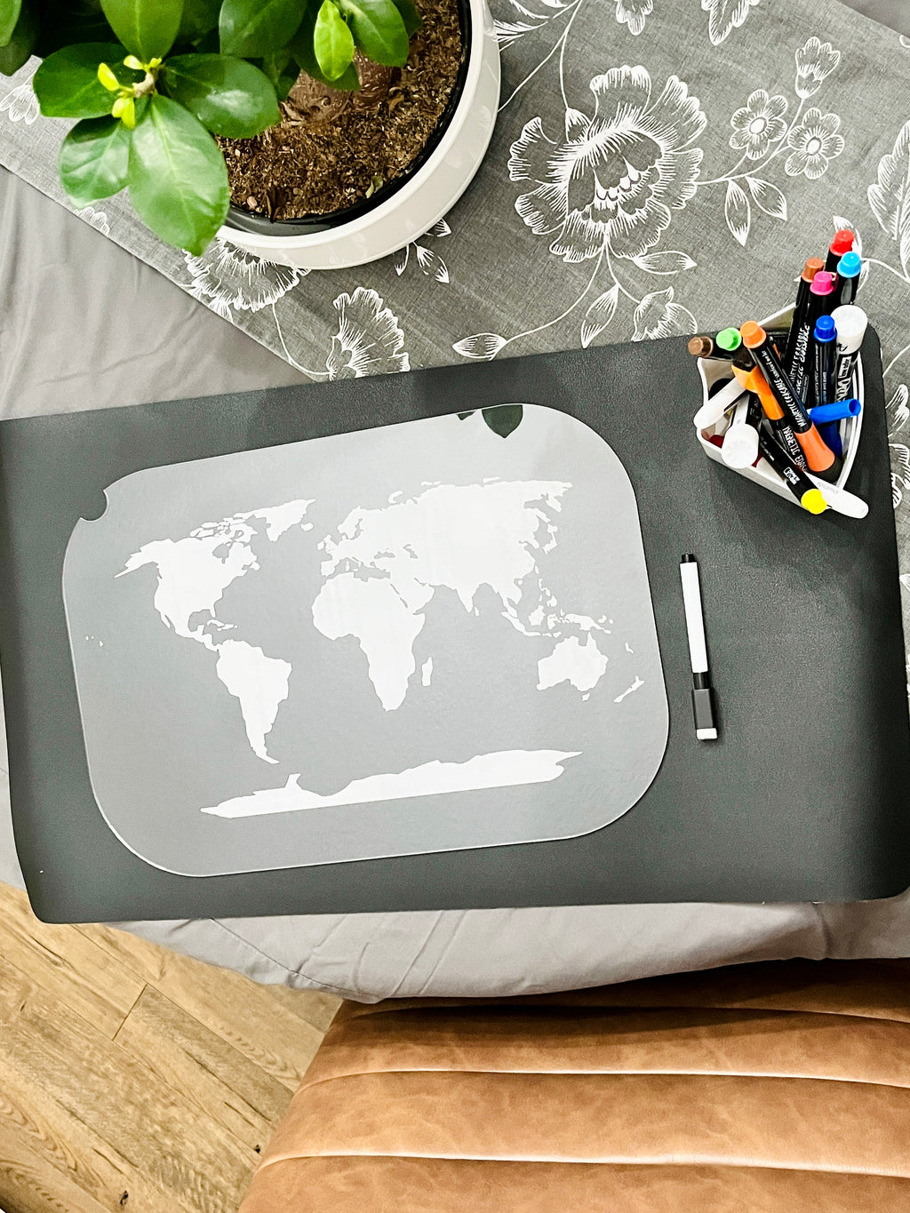 Gray placemat with a white world map outline beside a black dry erase marker and a container of colorful markers on a table