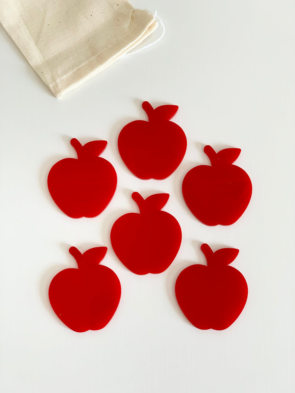 Six solid red apple-shaped cutouts arranged on a white surface next to a beige drawstring bag