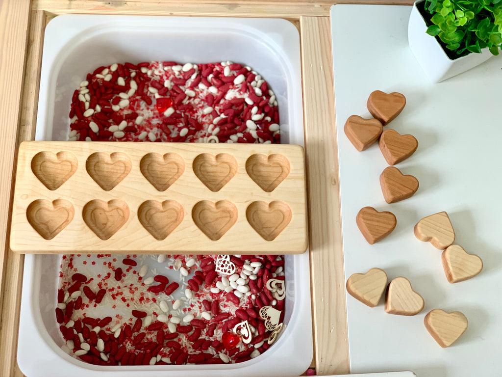 Wooden heart-shaped tray on white container filled with red and white rice, beans, and heart decorations, with wooden hearts nearby