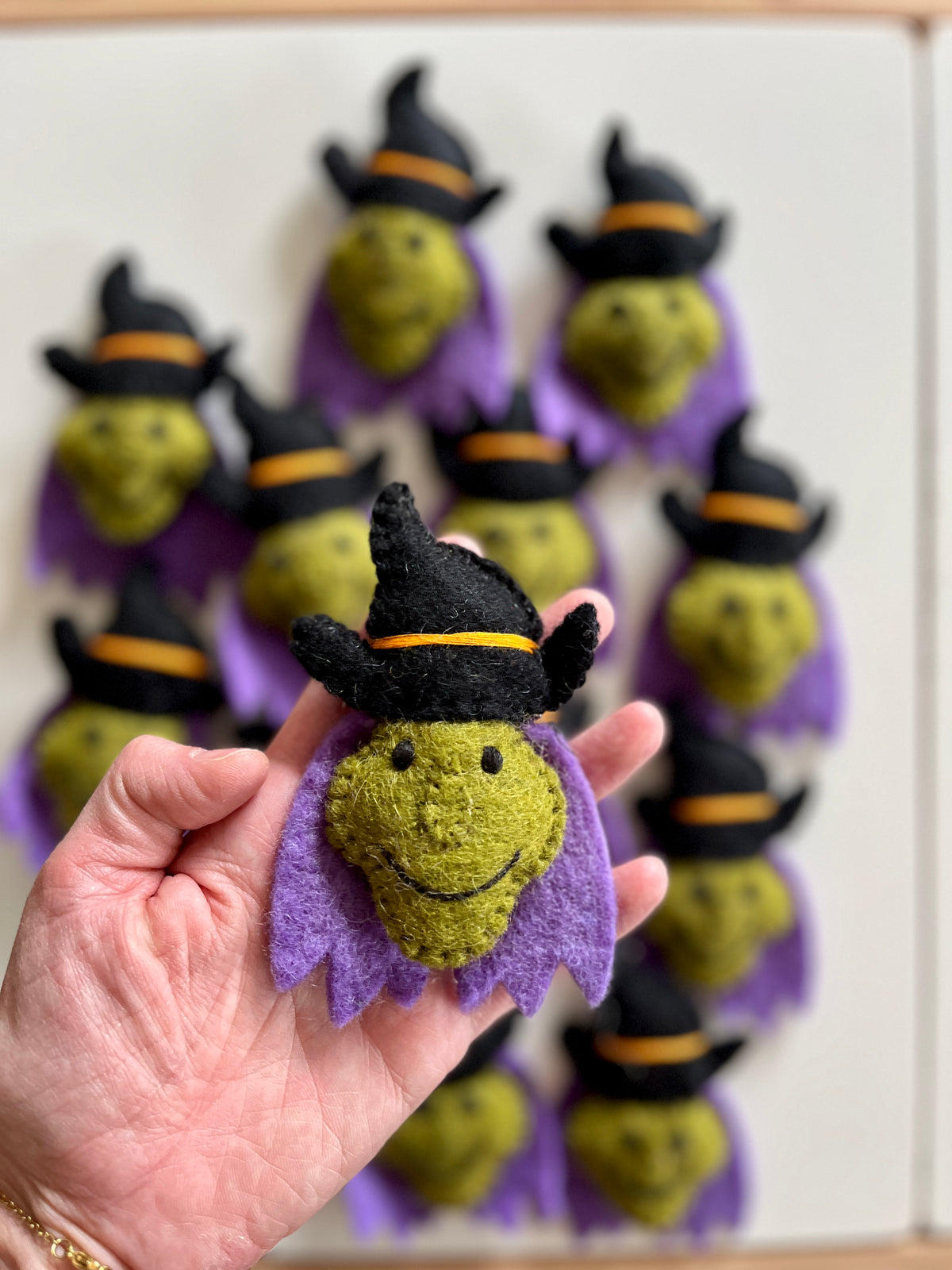 Hand holding a small green felt witch face with black hat and purple collar, with more similar witches blurred in background