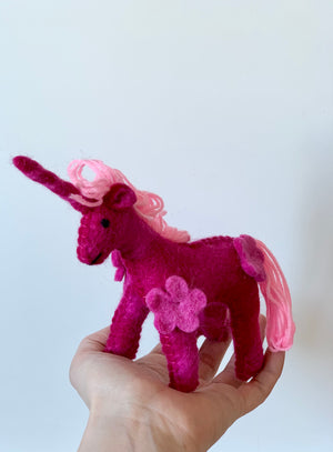 Hand holding a deep pink felt unicorn with light pink yarn mane, tail, and flower detail against a plain background