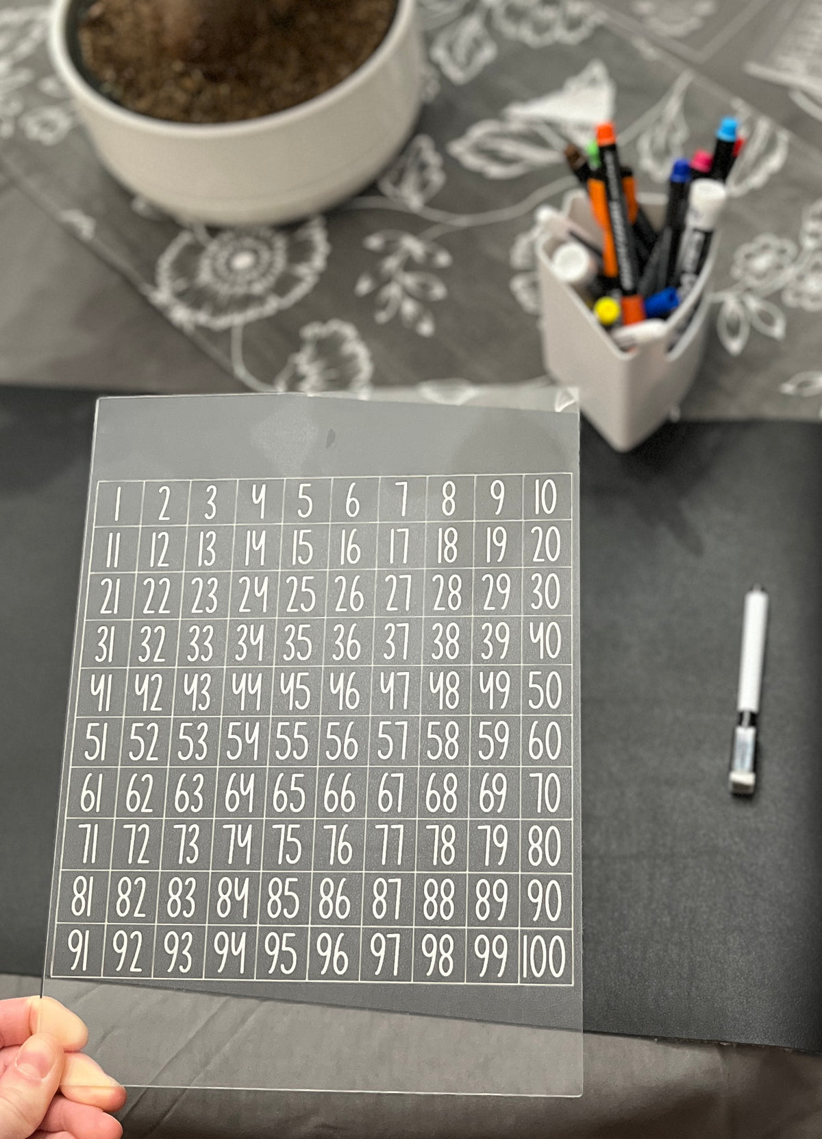 Hand holding transparent acrylic sheet with numbered grid 1 to 100 over a dark table with markers and plant nearby
