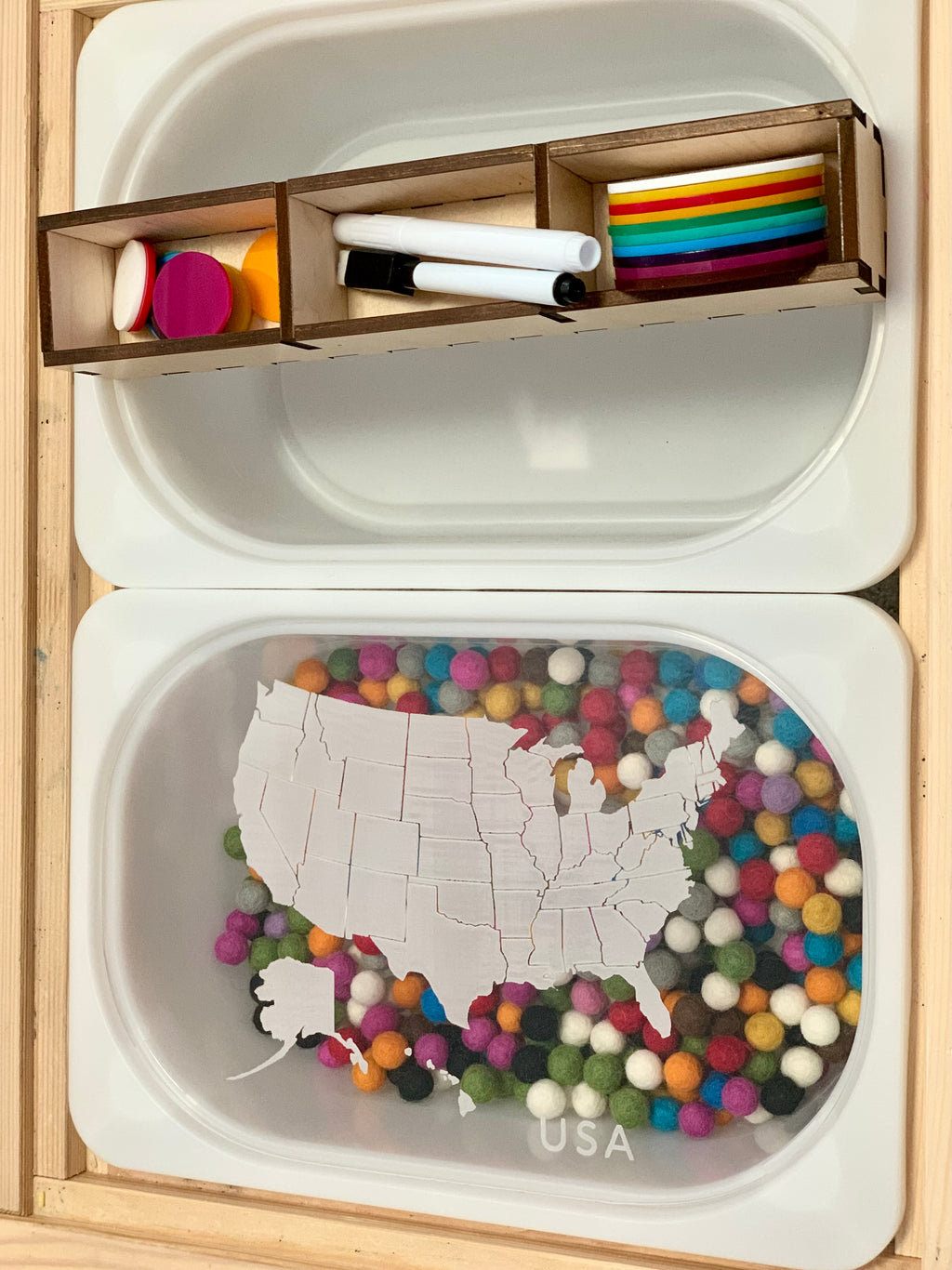 White tray with a puzzle map of the USA over colorful pom-poms and a wooden organizer holding markers and tokens above it
