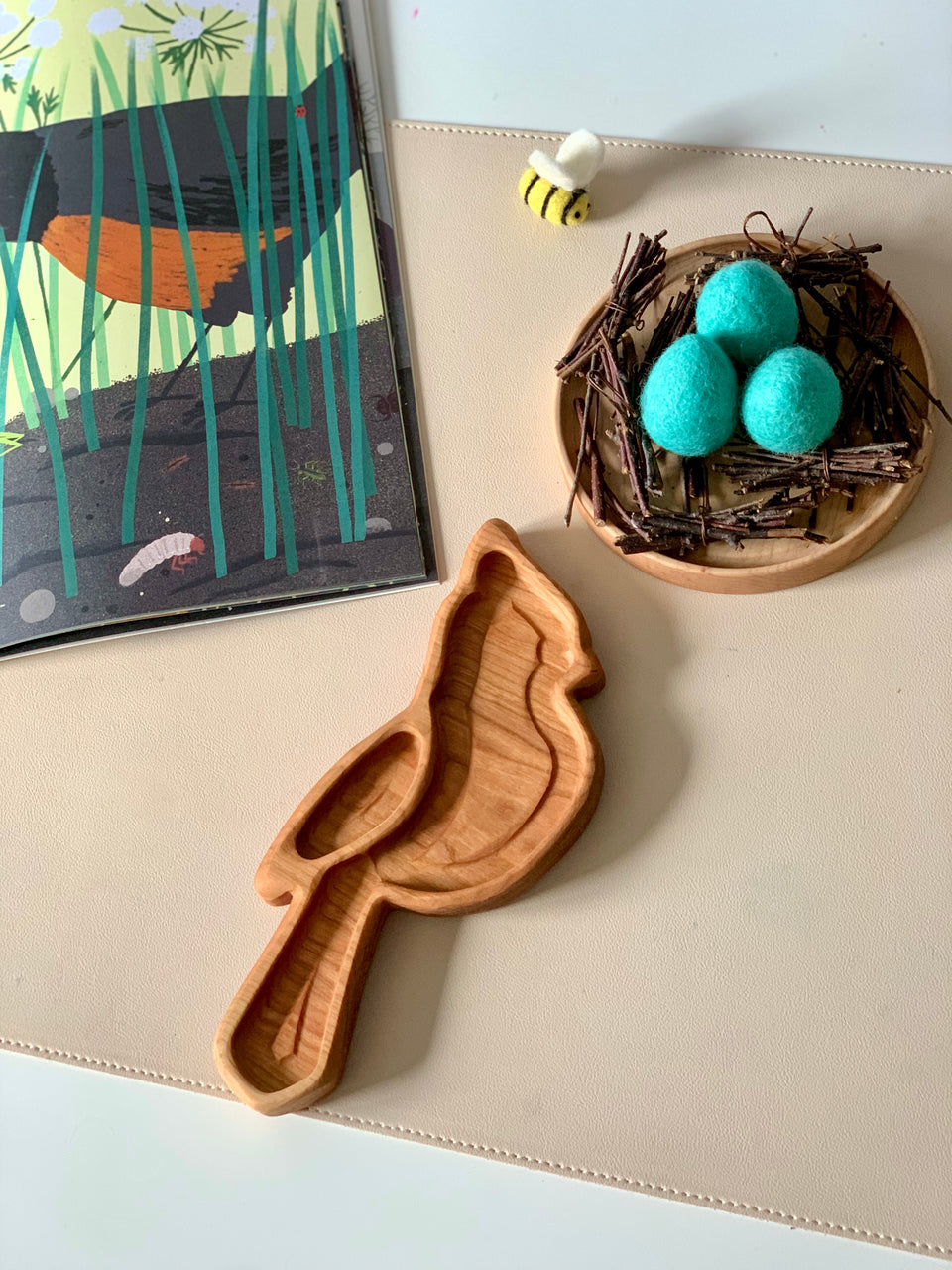 Wooden bird-shaped tray next to a small nest with three blue wool eggs and a felt bee on a beige surface