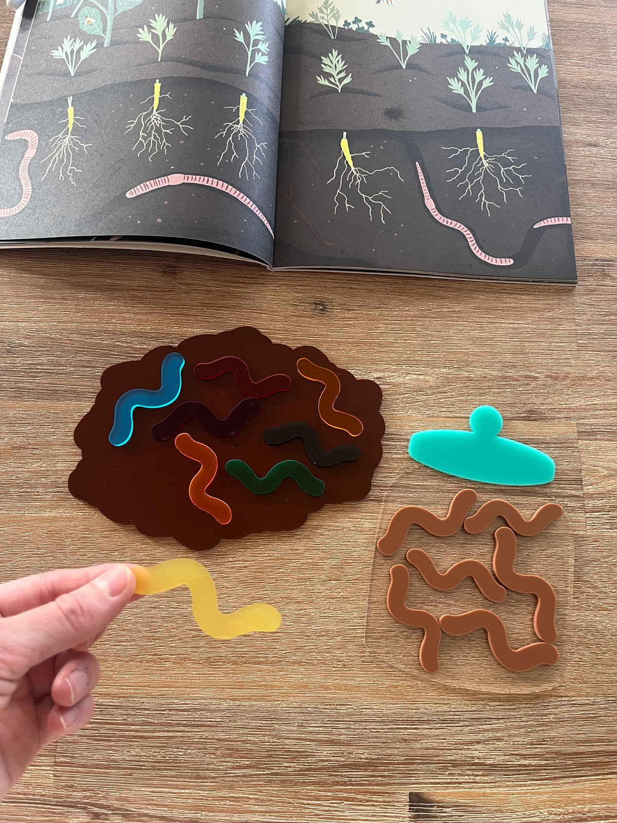 Little Dry Erasables - Worms with Dirt or Jar showing colorful translucent worm shapes on a brown dirt-shaped tray, a clear jar piece, and a hand holding a yellow worm, handcrafted homeschool education resources.