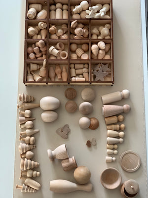 Assorted natural unfinished wooden shapes including peg dolls, trees, spheres, and stars organized in a compartment box and on table