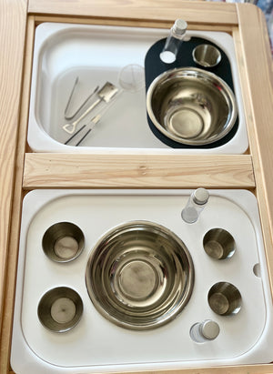 Wooden tray with stainless steel bowls, small cups, glass vials, and metal tongs on a white insert surface