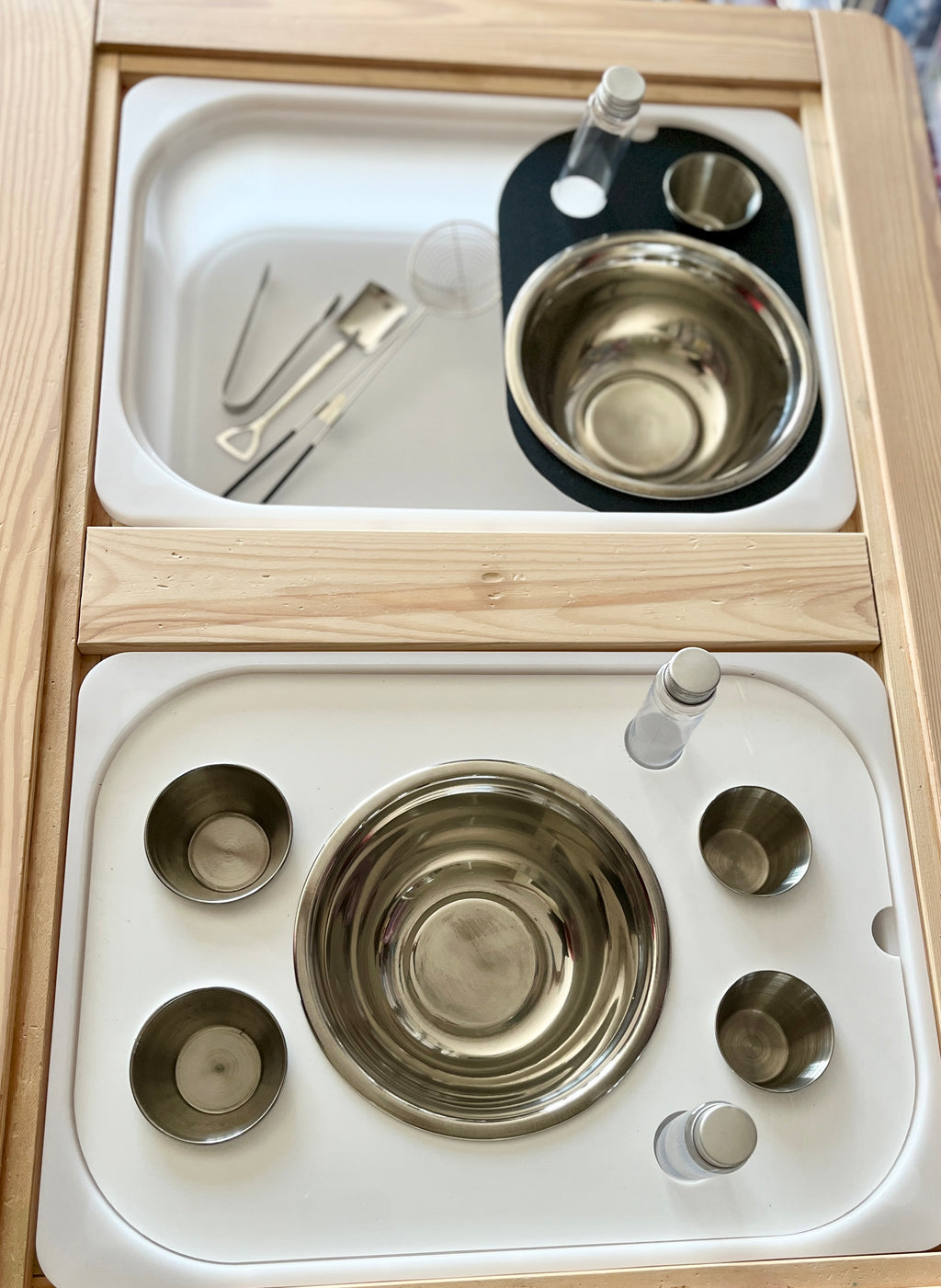 Wooden tray with stainless steel bowls, small cups, glass vials, and metal tongs on a white insert surface