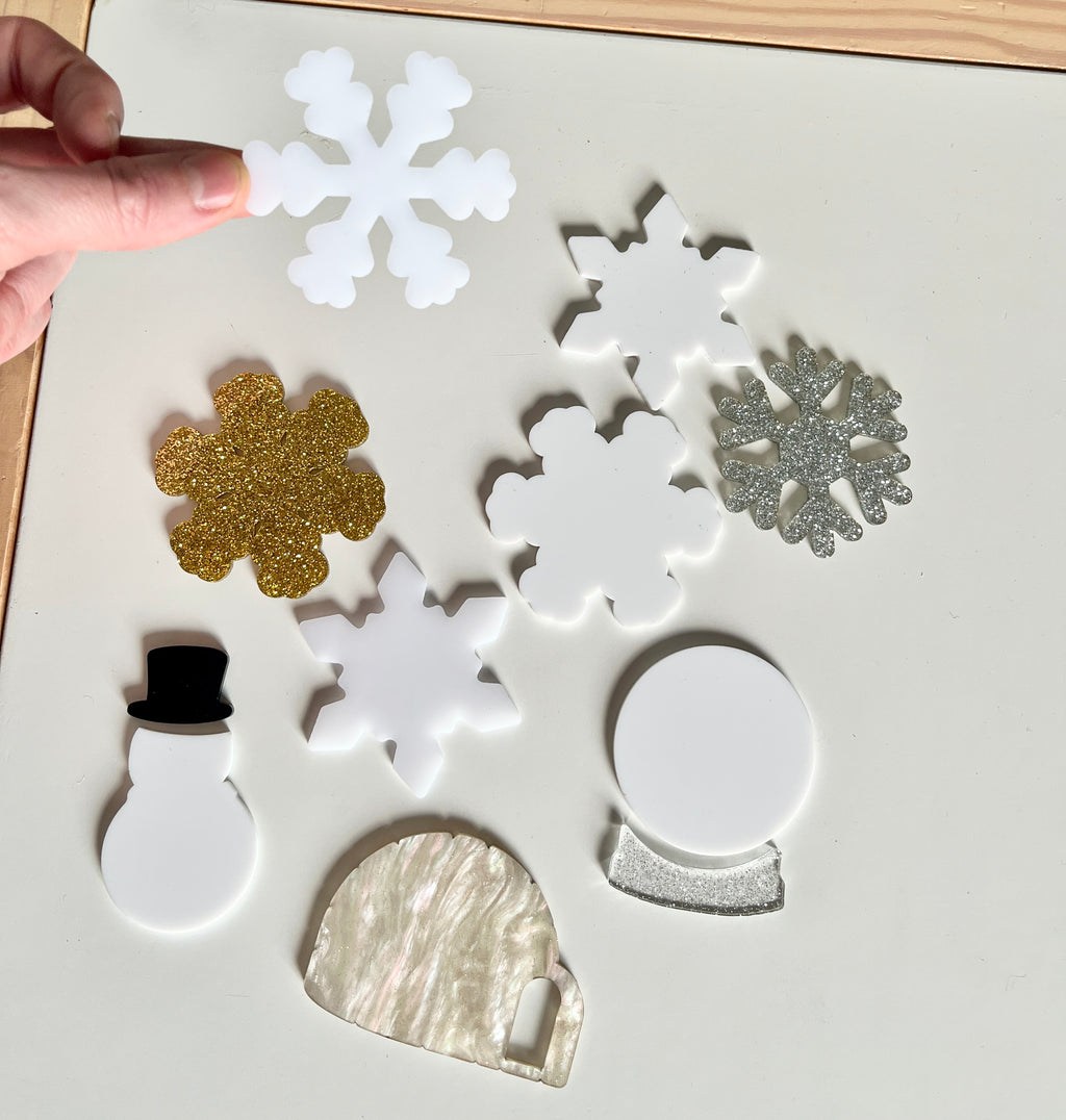 Hand holding white snowflake among various acrylic holiday shapes including gold, silver snowflakes, snowman, igloo, and snow globe on table.
