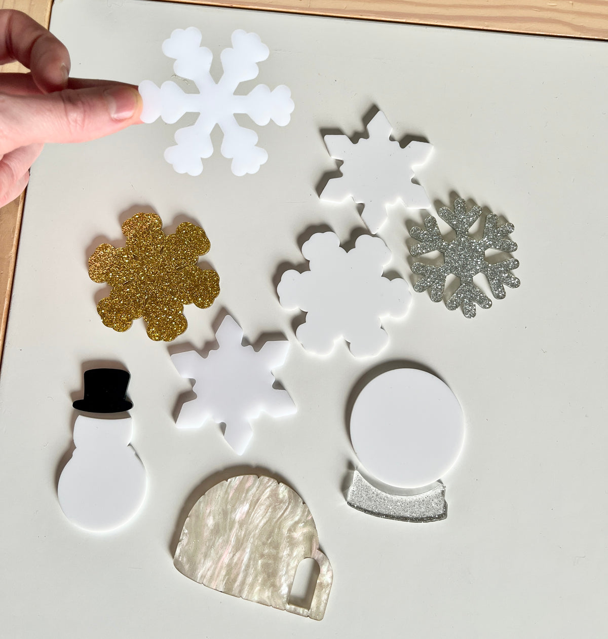 Hand holding white snowflake among various acrylic holiday shapes including gold, silver snowflakes, snowman, igloo, and snow globe on table.