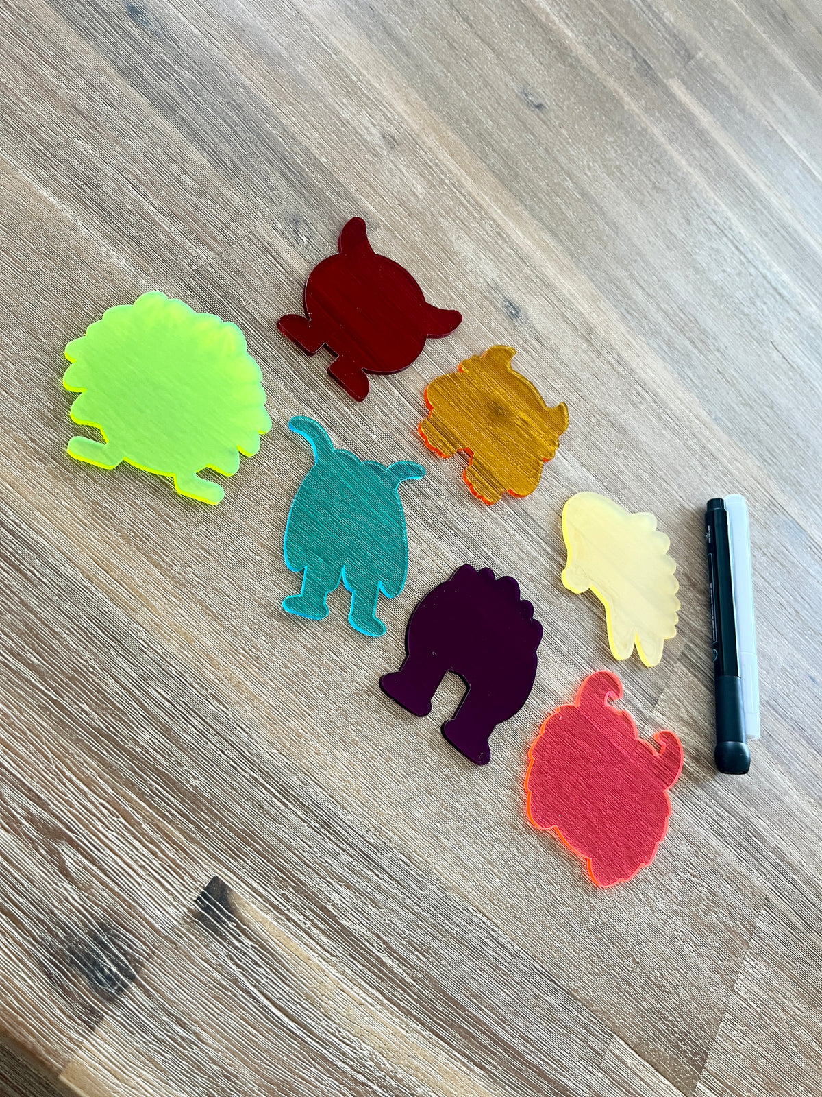 Seven colorful translucent monster-shaped pieces arranged on a wooden surface next to a black and white dry erase marker, featuring Little Dry Erasables — Translucent Monsters Set of 7, handcrafted homeschool education resources.