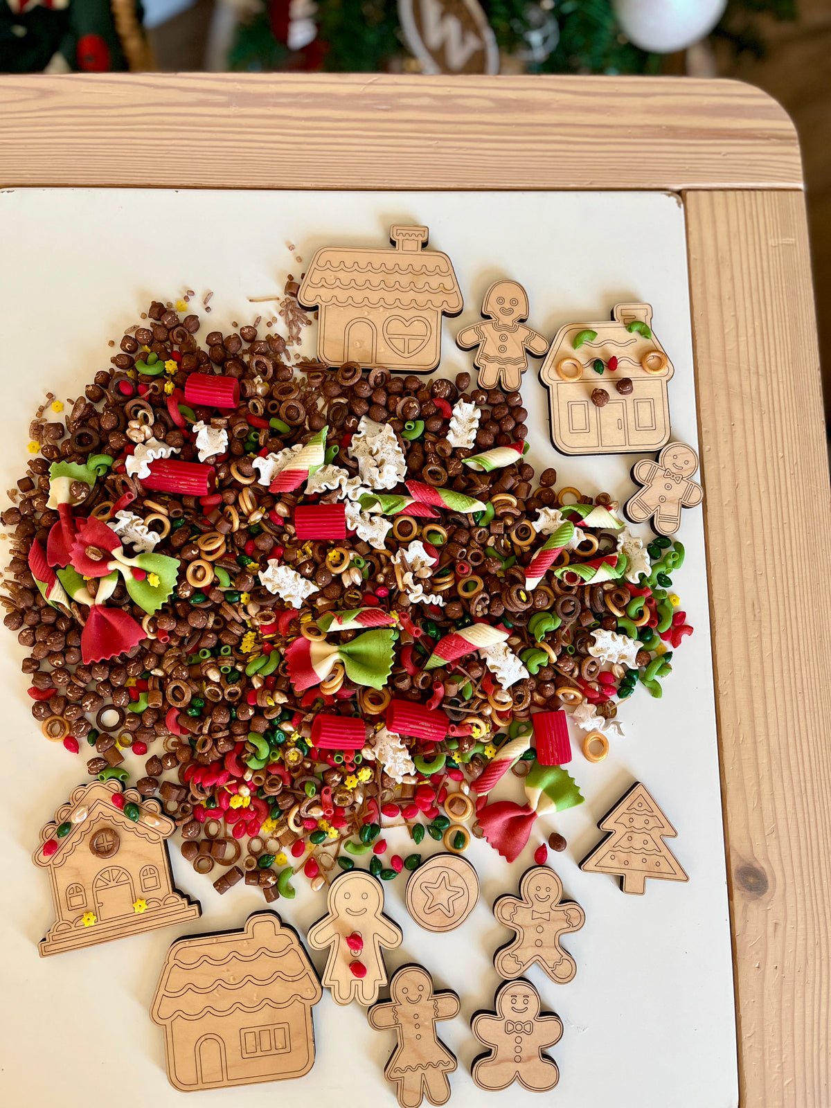 Assorted wooden holiday-themed cutouts surrounded by colorful pasta shapes and brown cereal pieces on a white surface
