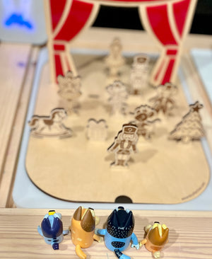 Four small animal figurines facing a wooden stage with holiday-themed laser-cut characters and red curtains