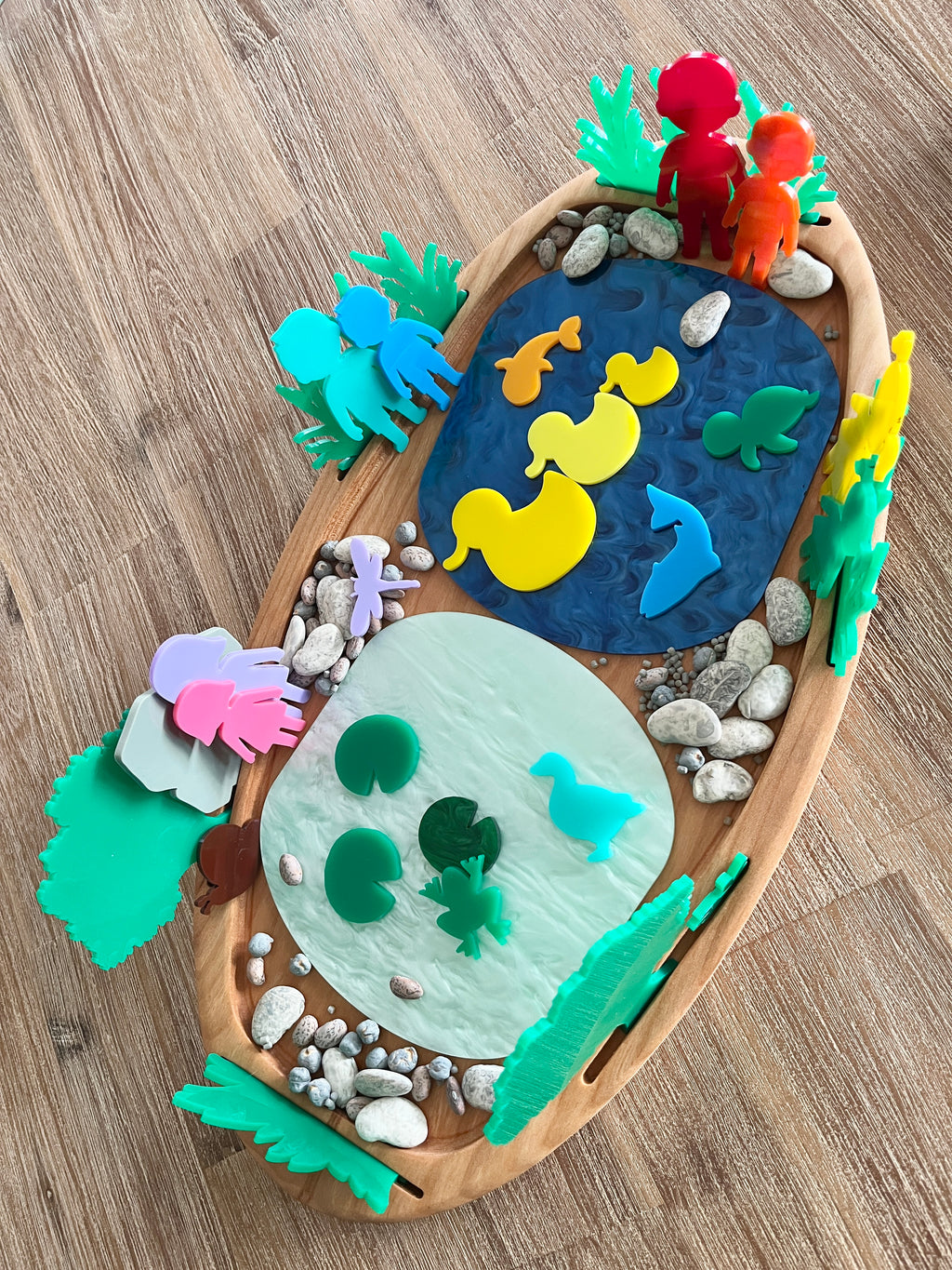 Multipurpose Oval Sensory Tray featuring a wooden tray with two textured sensory ponds in blue and white, colorful acrylic animal figures, natural stones, and green plant shapes, handcrafted, homeschool, education resources.