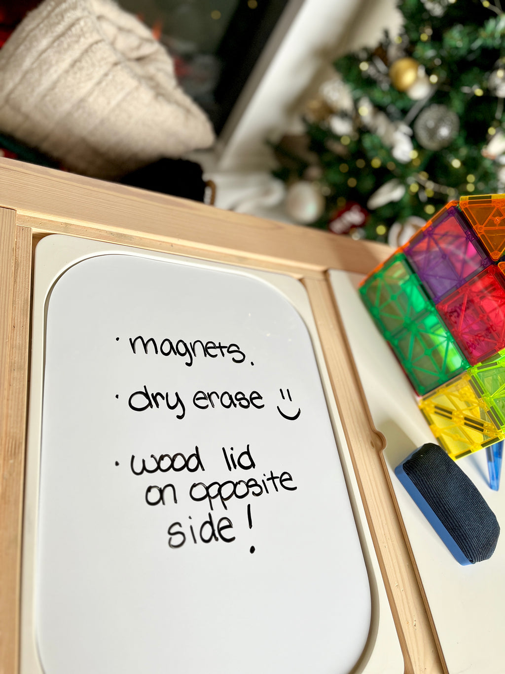 White dry erase board with handwritten text, wooden frame, colorful magnetic tiles, and blurred holiday tree background
