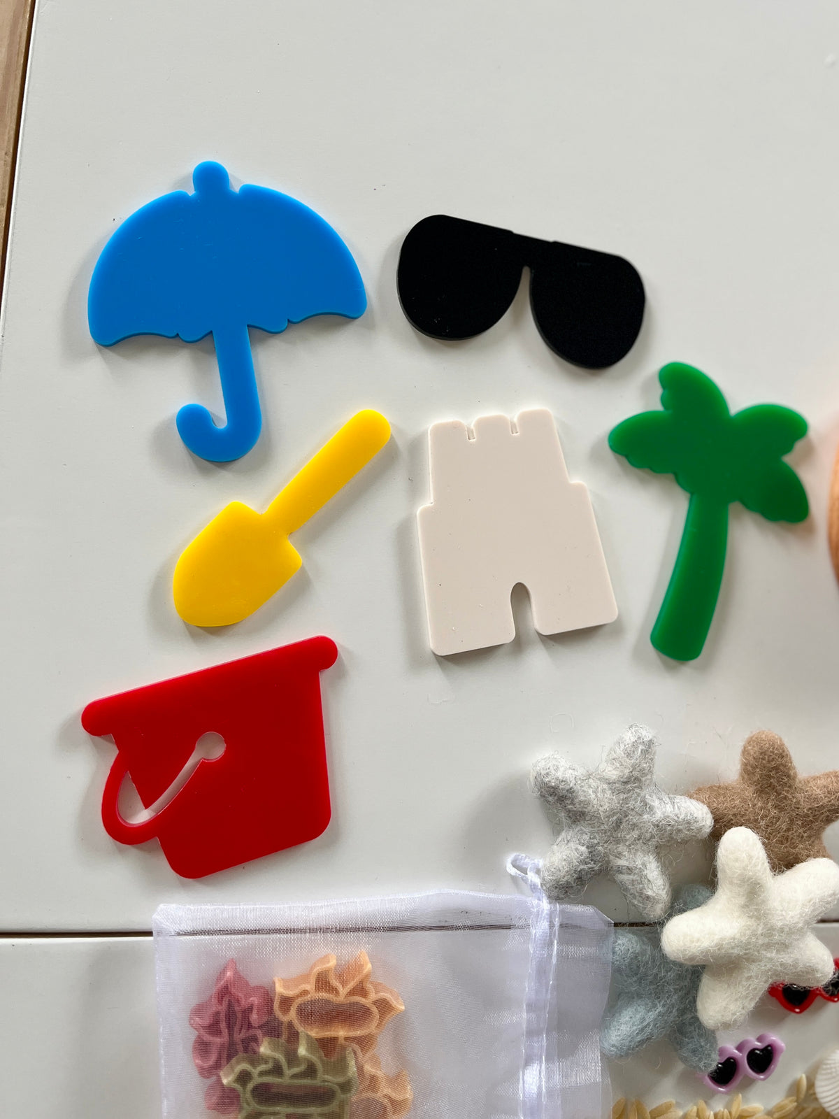 Colorful plastic shapes including umbrella, sunglasses, palm tree, shovel, bucket, and castle with felt starfish on white surface